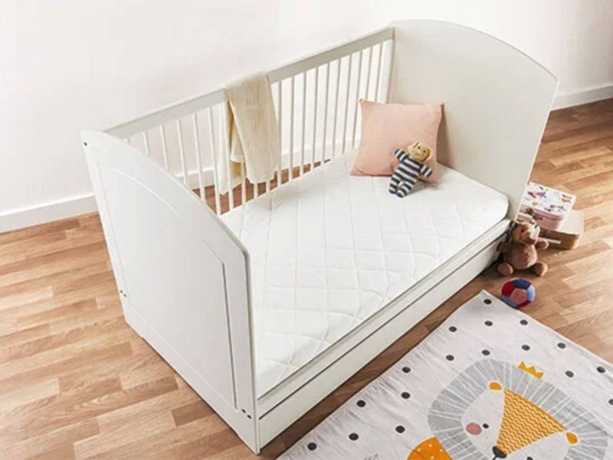 Lit bébé blanc avec matelas, oreiller et jouets sur un sol en bois.