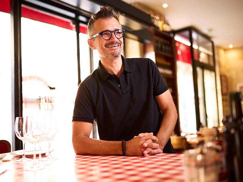 Homme souriant en polo noir, accoudé à une table à carreaux rouges et blancs dans un restaurant.
