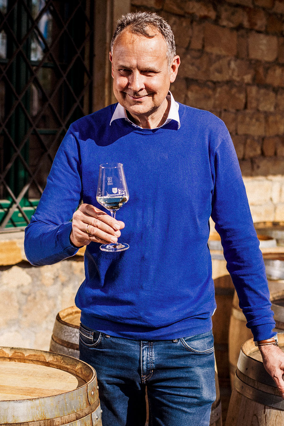 Lächelnder Mann in blauem Pullover und Jeans, hält ein Glas Weißwein zwischen Holzfässern.