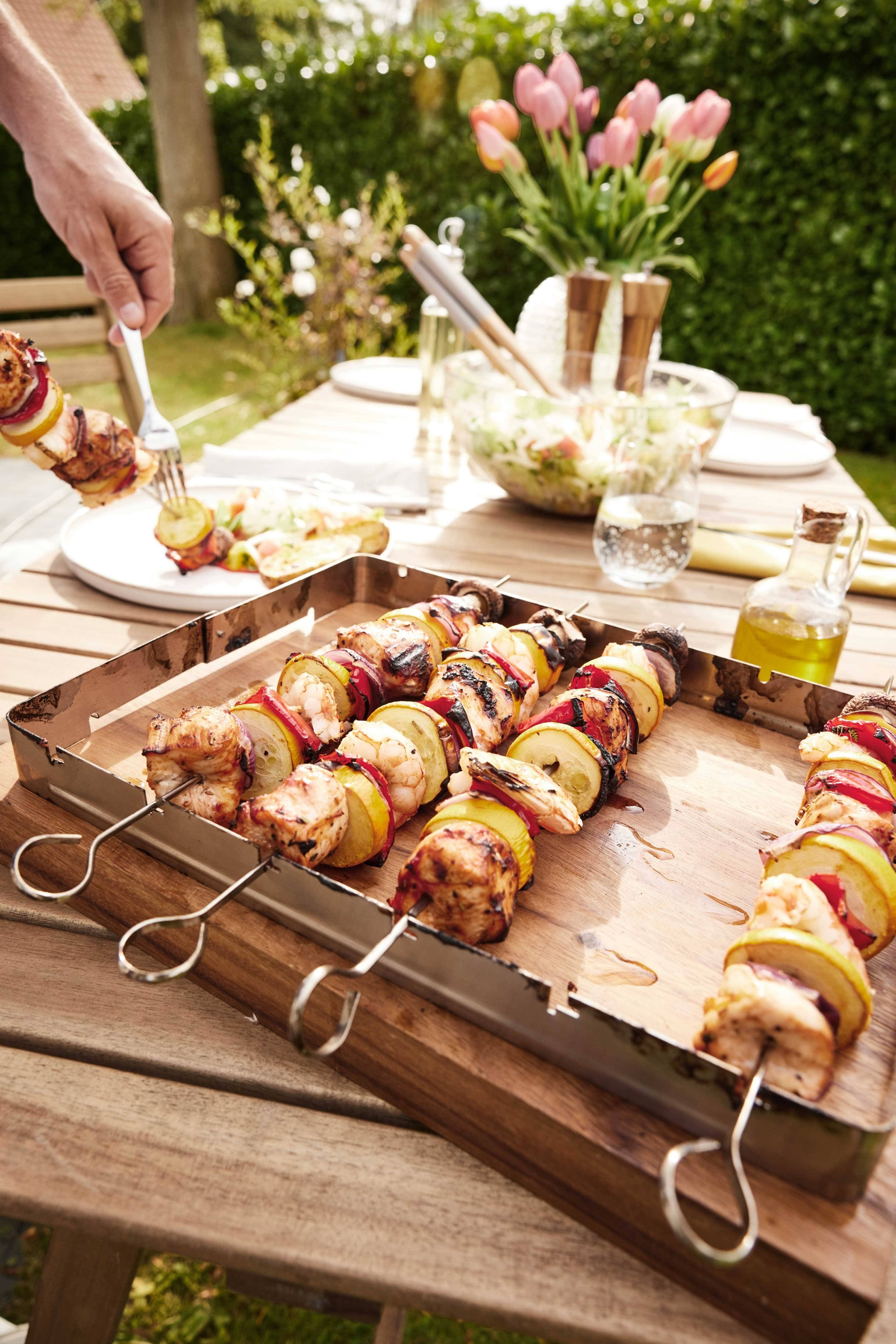 Brochettes de poulet et légumes sur une table en bois extérieure, avec salade et tulipes.