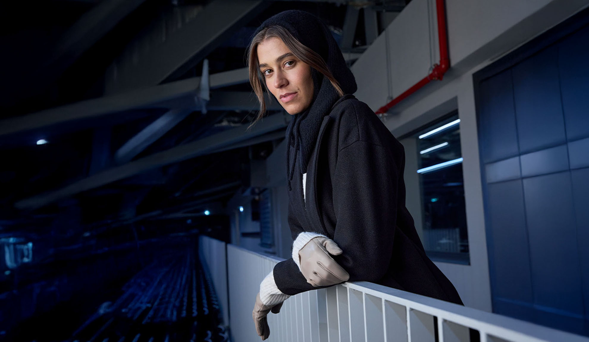 Femme en manteau noir et écharpe à capuche, appuyée sur une balustrade dans un cadre moderne.