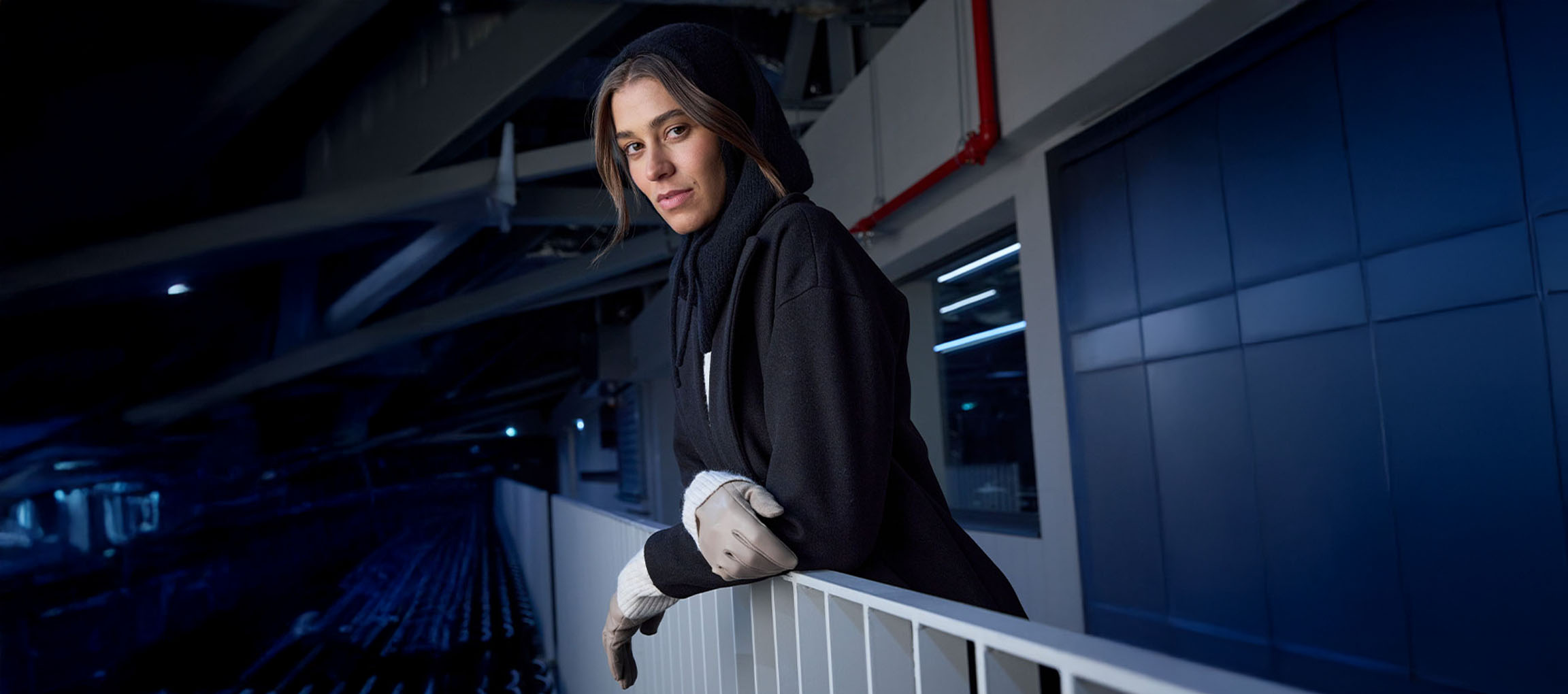 Femme en manteau noir et gants appuyée sur une balustrade en milieu urbain.