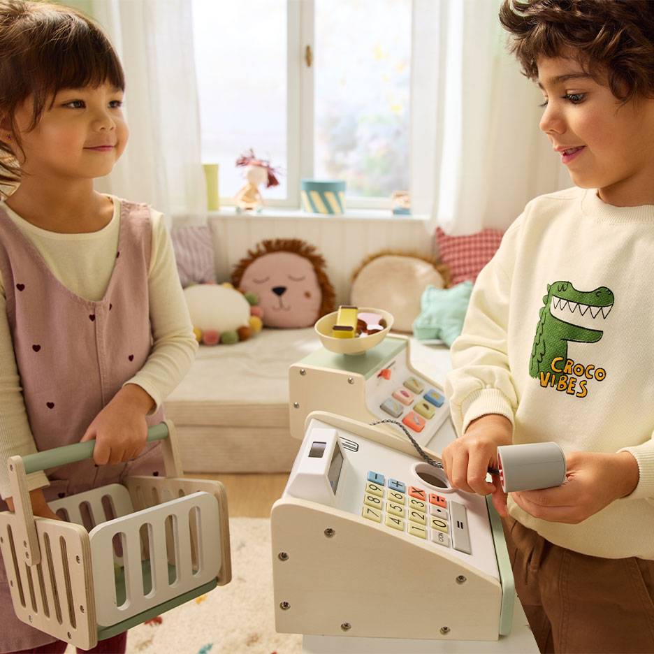 Enfants jouant à la marchande; garçon utilisant une caisse enregistreuse, fille portant un panier.
