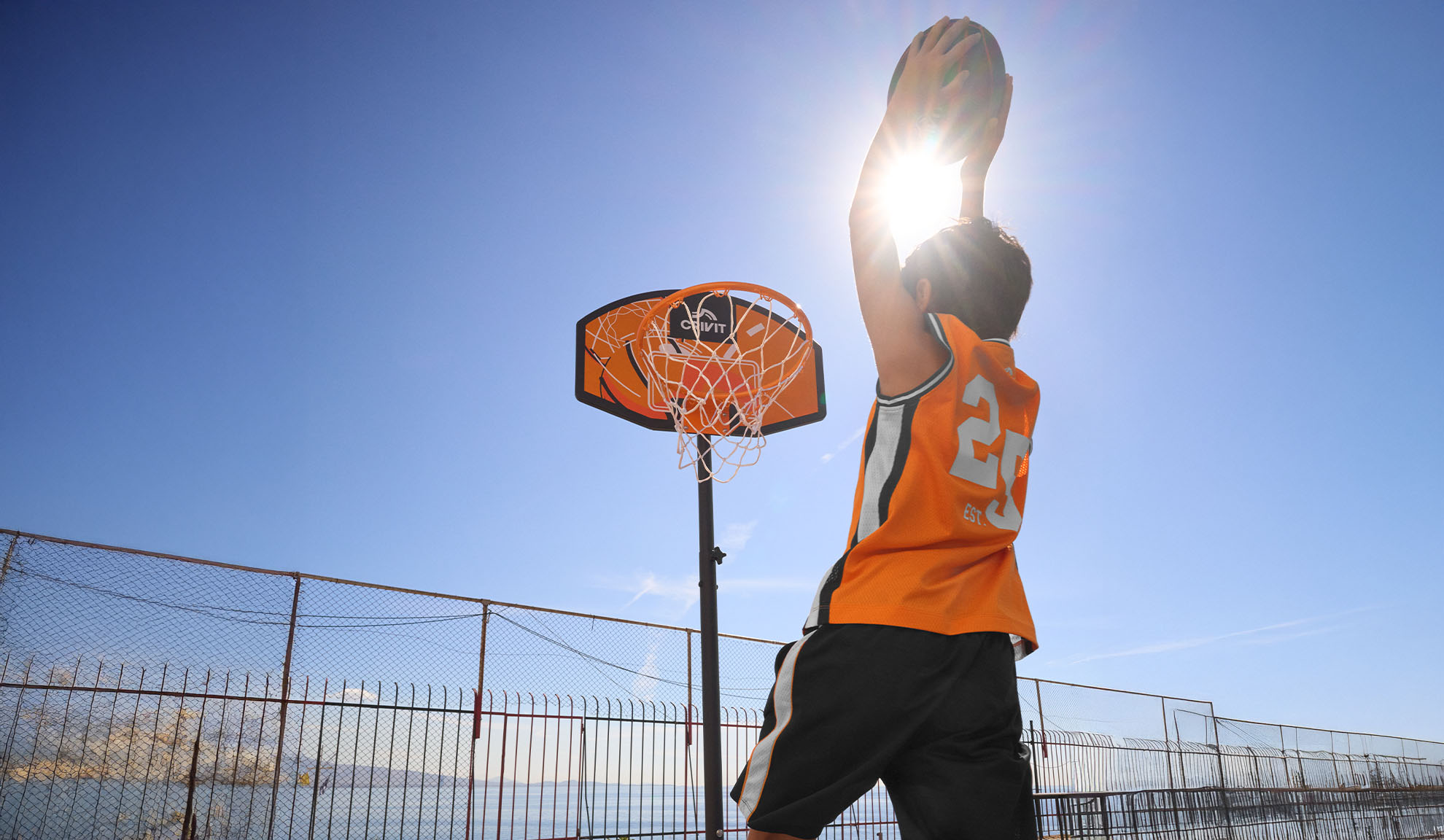 Garçon en maillot de basket tirant au panier en extérieur sous un soleil éclatant.