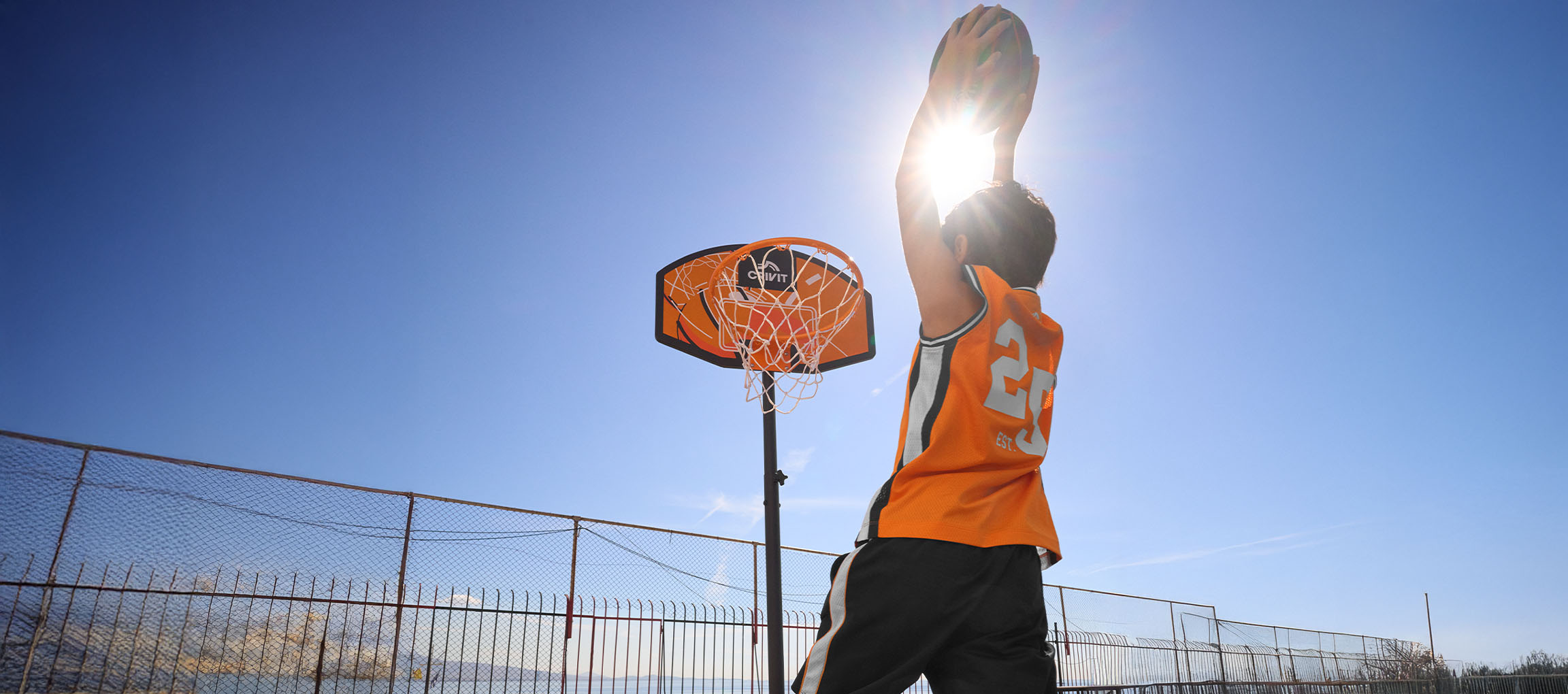 Garçon en maillot de basket orange lançant un ballon dans le panier par une journée ensoleillée.