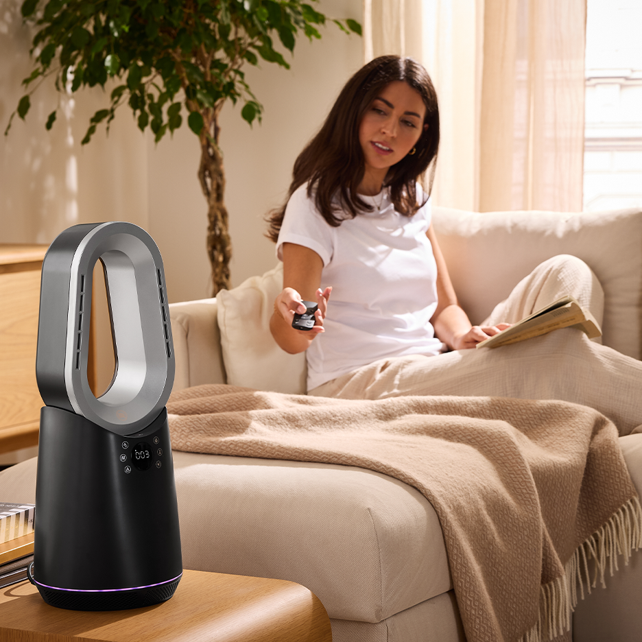 Eine Frau auf einem Sofa steuert mit einer Fernbedienung einen schwarz-silbernen, blattlosen Ventilator.