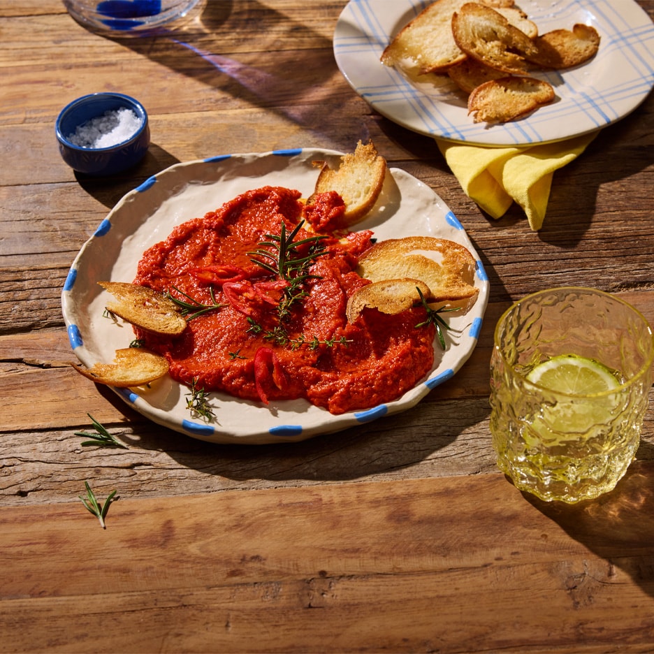 Roter Paprika-Dip mit Rosmarin und geröstetem Brot auf einem rustikalen Holztisch.