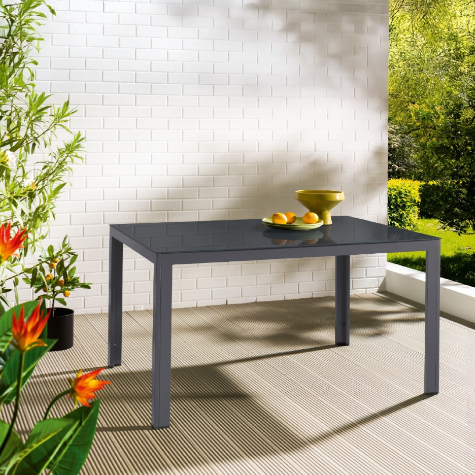 Grauer Gartentisch mit Glasplatte, dekoriert mit Pflanzen und Obst auf einer Terrasse.