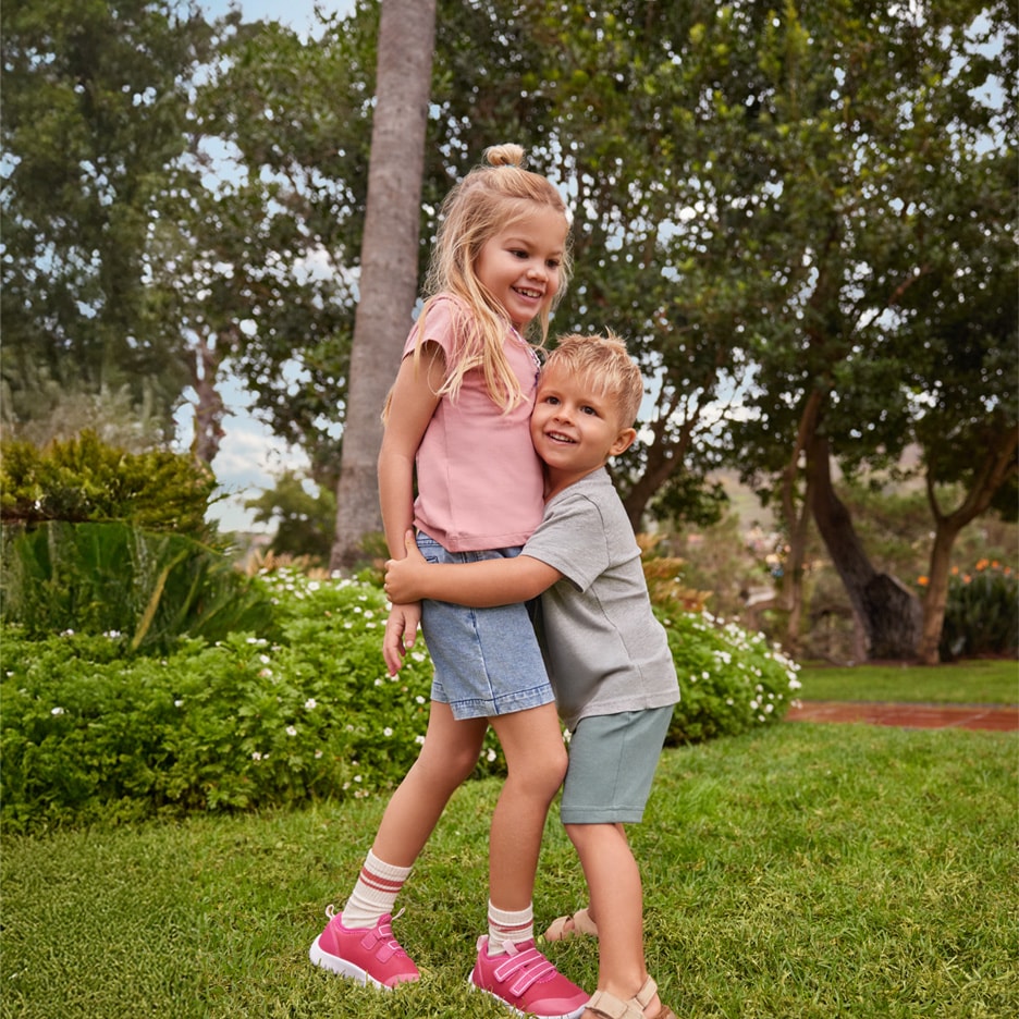 Zwei lächelnde Kinder in Sommerkleidung, das Mädchen trägt Jeansshorts und pinke Turnschuhe.