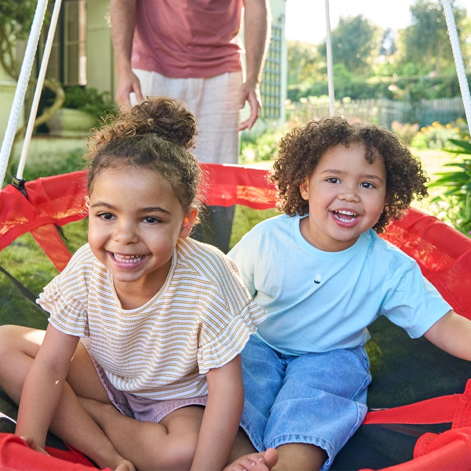Zwei lächelnde Kinder spielen draußen auf einer roten Schaukel.