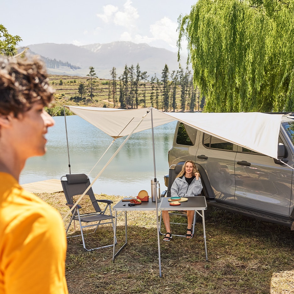 Un auvent de voiture et une table de camping avec une femme assise près d'un lac.
