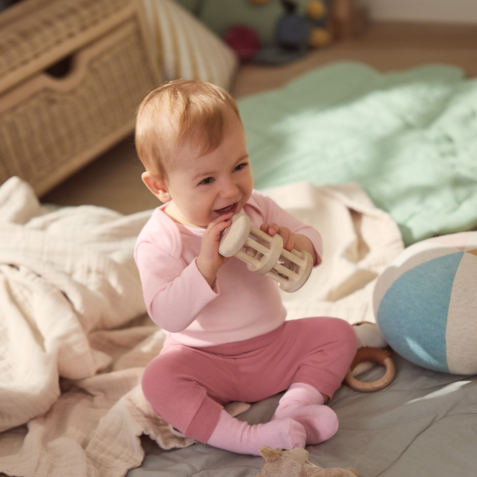 Glückliches Baby in rosa Kleidung spielt mit Holzrassel auf einer Decke.