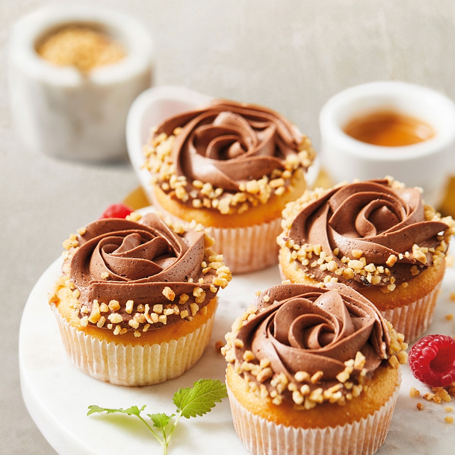 Vier Vanille-Cupcakes mit rosenförmiger Schokoladenglasur und gehackten Haselnüssen.