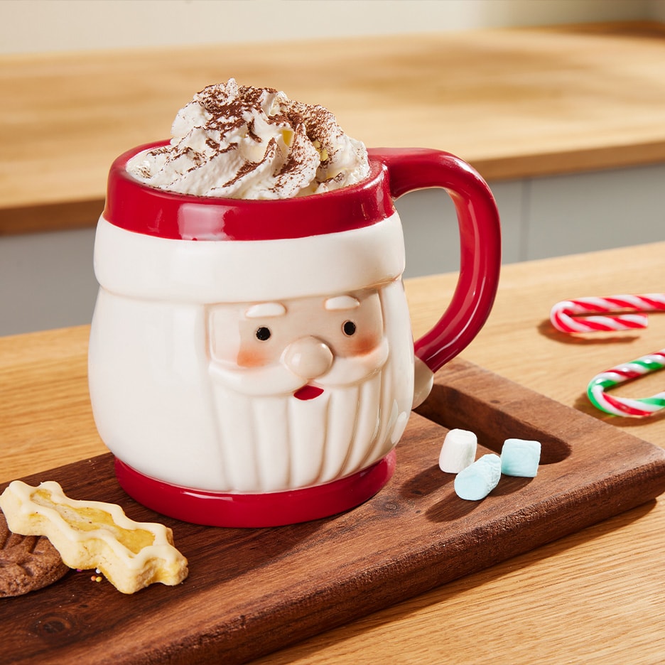 Weihnachtsmann-Tasse mit heißer Schokolade, Schlagsahne und Kakaopulver, auf einem Holztablett mit Keksen, Marshmallows und Zuckerstangen.
