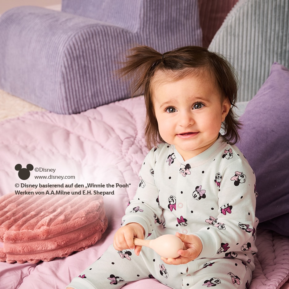 Lächelndes Baby in Disney Minnie Maus Schlafanzug, spielt mit einer Rassel.