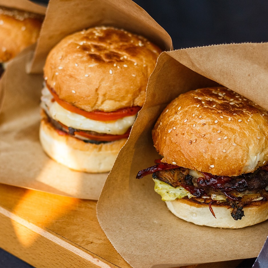 Zwei Burger in Papiertüten auf einem Holzbrett, einer mit Tomate und Käse, der andere mit Pilzen.