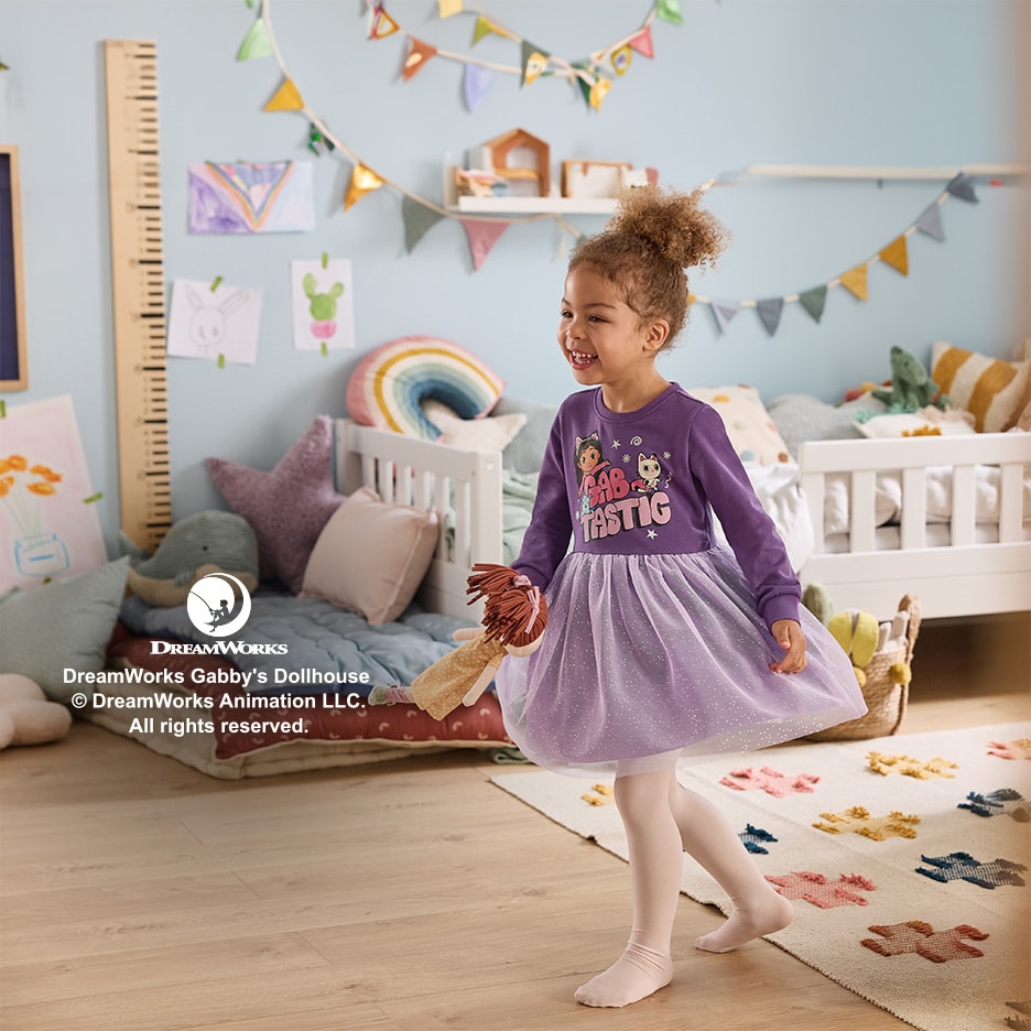 Lachendes Mädchen in lila Gabby's Dollhouse Kleid mit Puppe in einem Kinderzimmer.