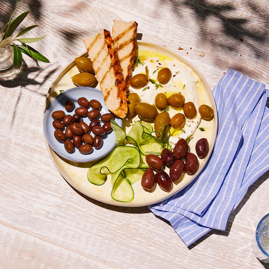 Teller mit gemischten Oliven, Tzatziki, Gurken und geröstetem Brot auf einem Holztisch.