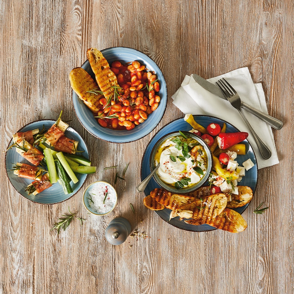 Holztisch mit drei Vorspeisenplatten: gebackene Bohnen, Schinken-Käse-Spieße mit Gemüse und geröstetes Brot mit Dip.