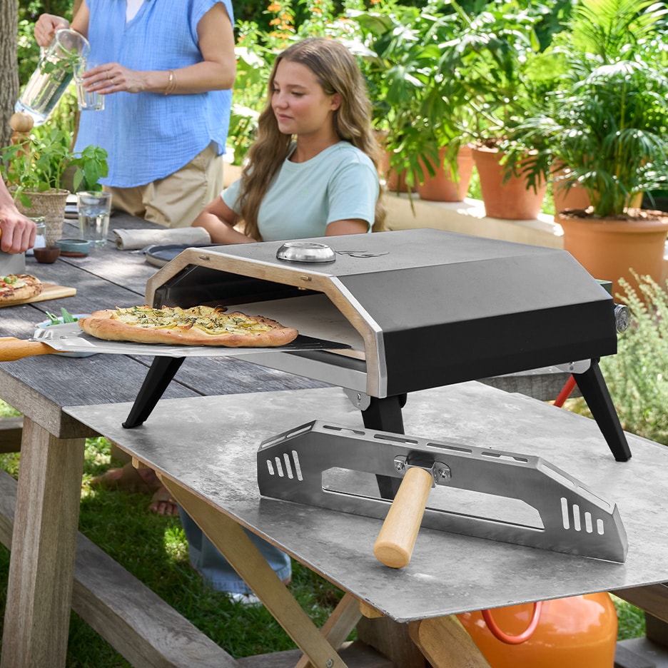 Outdoor-Pizzaofen mit frisch gebackener Pizza auf einem Holztisch, umgeben von Personen.