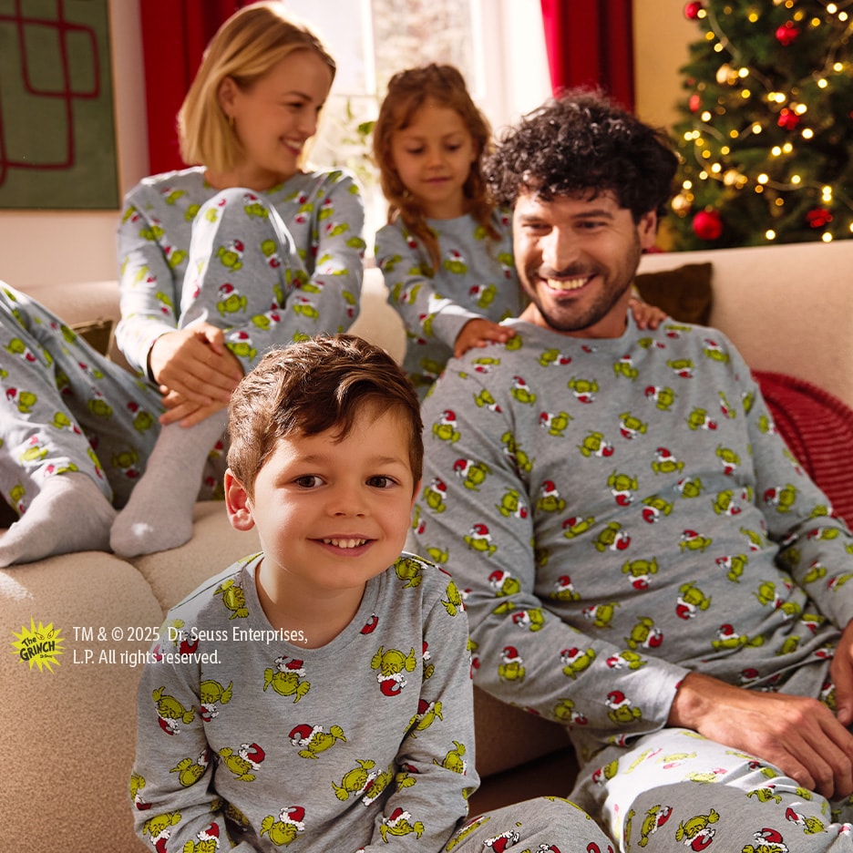 Familie in passenden Grinch-Weihnachtspyjamas, lächelnd auf einem Sofa mit Weihnachtsbaum.