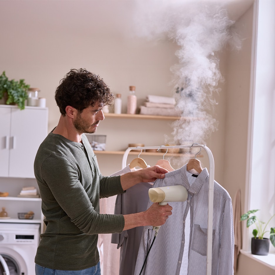 Mann dampft Hemd mit Handdampfglätter auf Kleiderständer in der Waschküche.