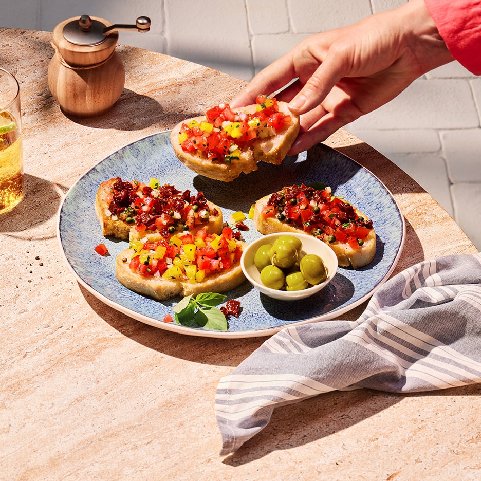Eine Hand nimmt Bruschetta mit Tomaten und Paprika von einem Teller mit Oliven und einem Getränk.