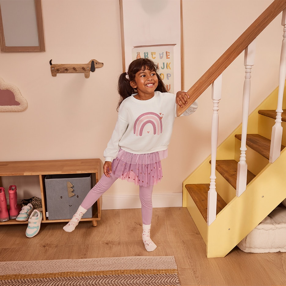 Ein lächelndes kleines Mädchen trägt einen weißen Pullover mit Regenbogenmotiv und einen rosa Tüllrock.