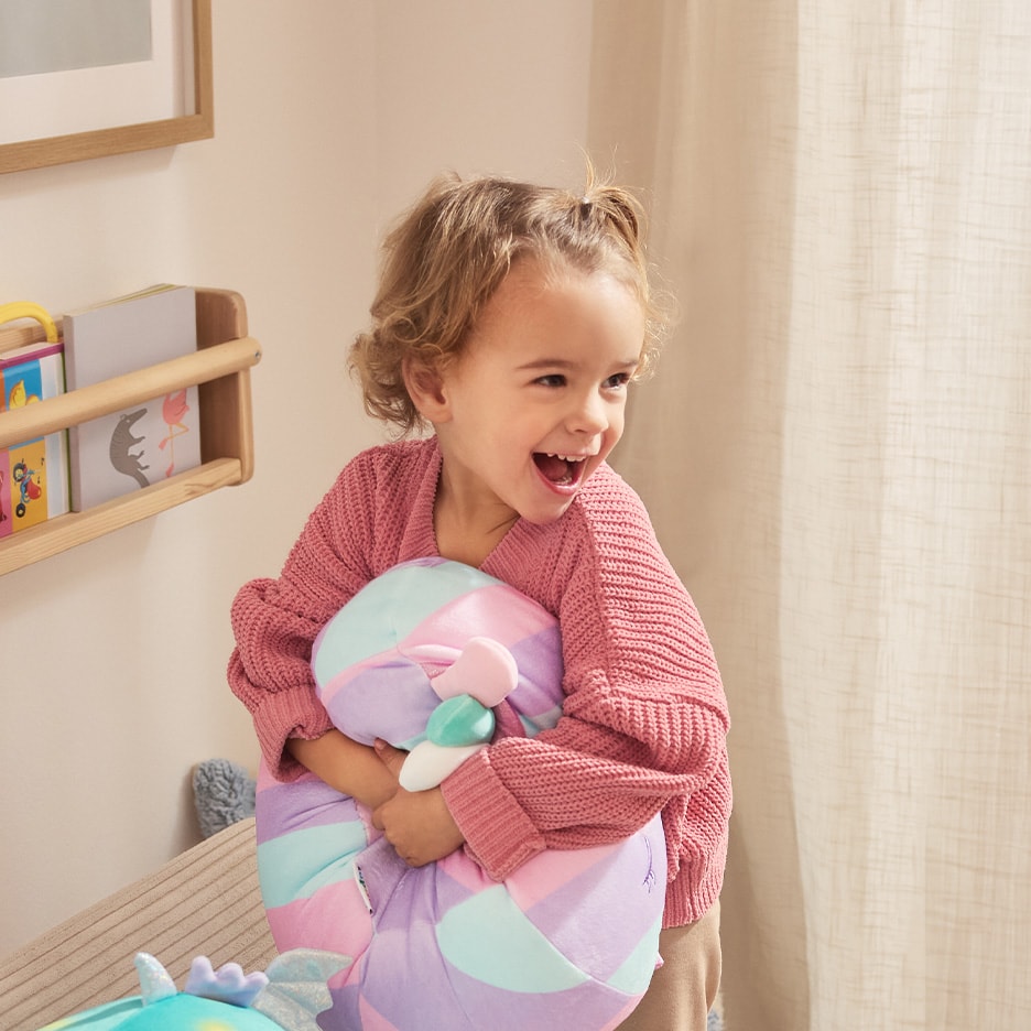 Petite fille souriante en pull rose, serrant un coussin licorne, dans une chambre avec étagère.