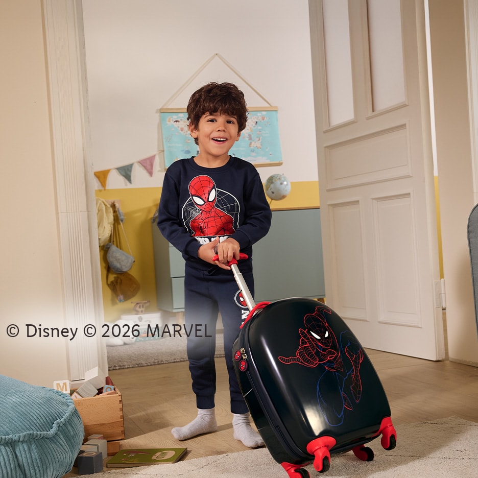 Lächelndes Kind im Superhelden-Schlafanzug zieht einen passenden Rollkoffer.
