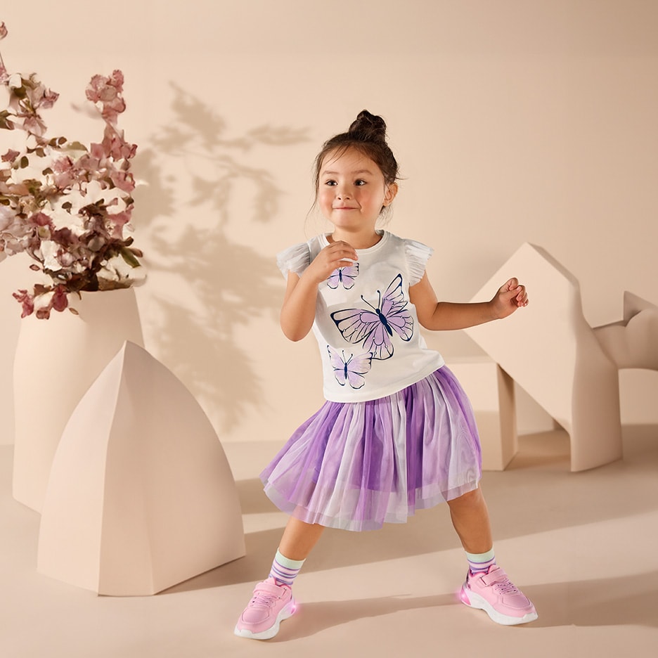 Kleines Mädchen in Schmetterlings-T-Shirt und lila Tüllrock, lächelnd und tanzend.