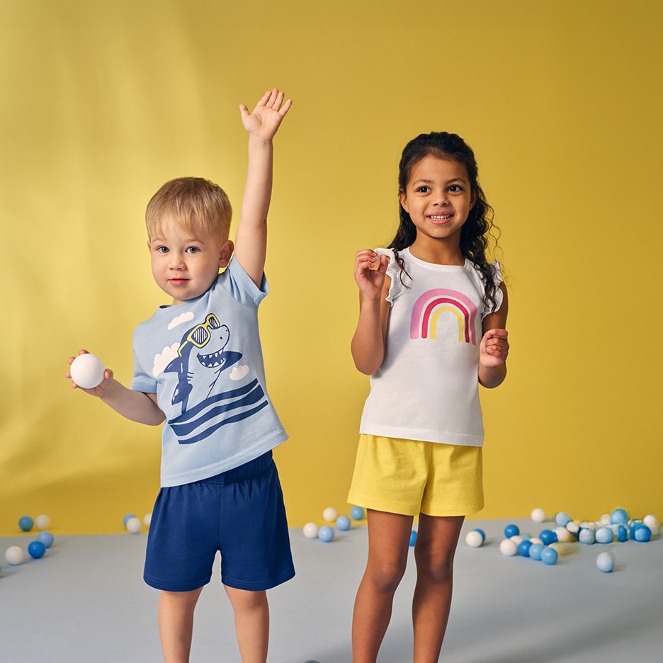 Kinder in Sommer-T-Shirts und Shorts, Junge trägt Haifisch-T-Shirt, Mädchen trägt Regenbogen-T-Shirt.