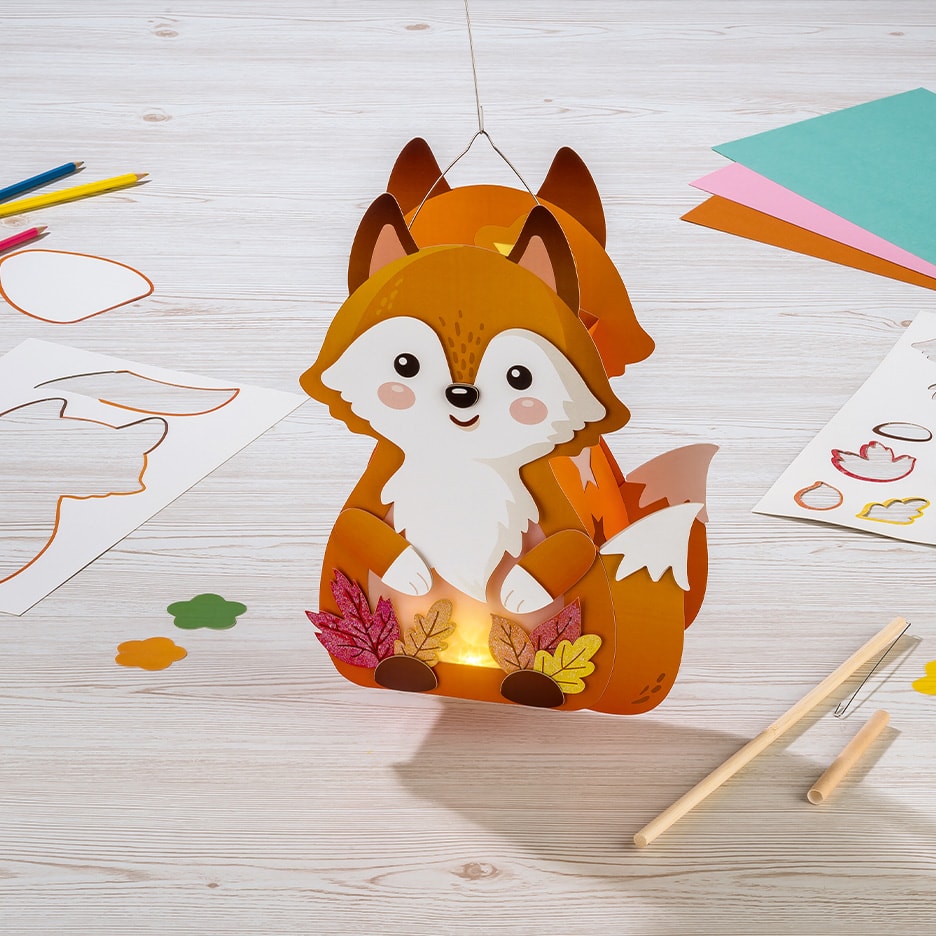 Papierfuchs-Laterne mit Bastelmaterialien auf einem Holztisch.