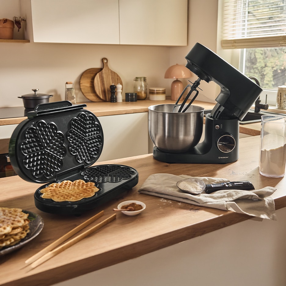 Schwarzes Waffeleisen und Küchenmaschine auf einer Holzküchenarbeitsplatte mit Waffeln.