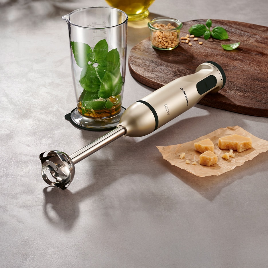 Ein goldener Stabmixer mit Pesto-Zutaten auf einer Arbeitsplatte.