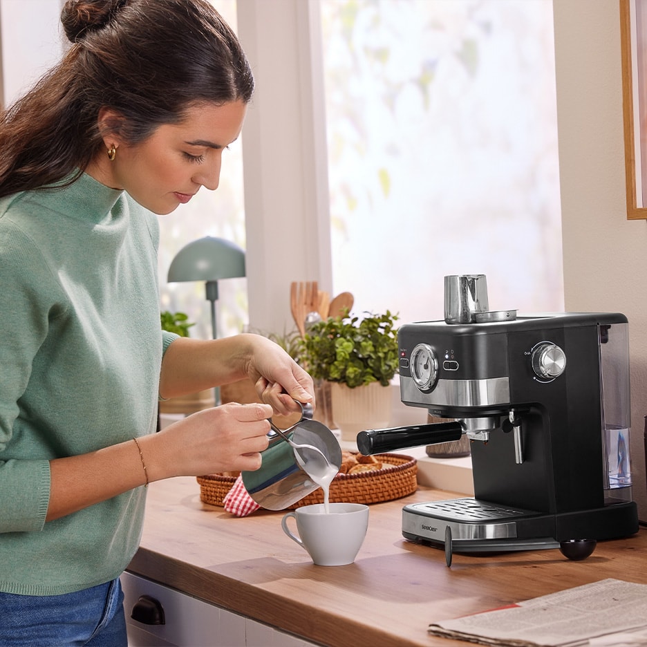 Frau gießt aufgeschäumte Milch aus einem Krug in eine Tasse, neben einer Silvercrest Kaffeemaschine.