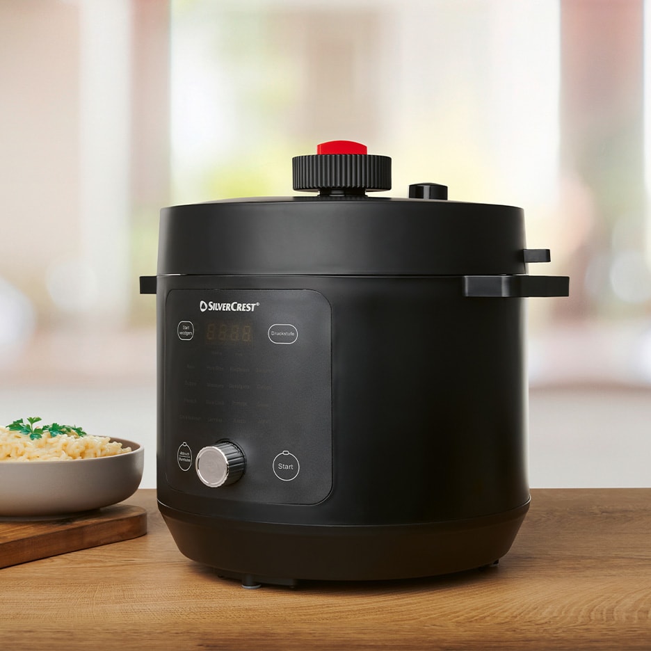 Schwarzer Silvercrest Multifunktions-Elektro-Dampfkochtopf auf einem Holztisch mit einer Schüssel Nudeln.