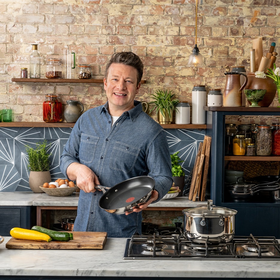 Ein lächelnder Koch hält eine Pfanne in einer rustikalen Küche mit Kochgeschirr und Gemüse.