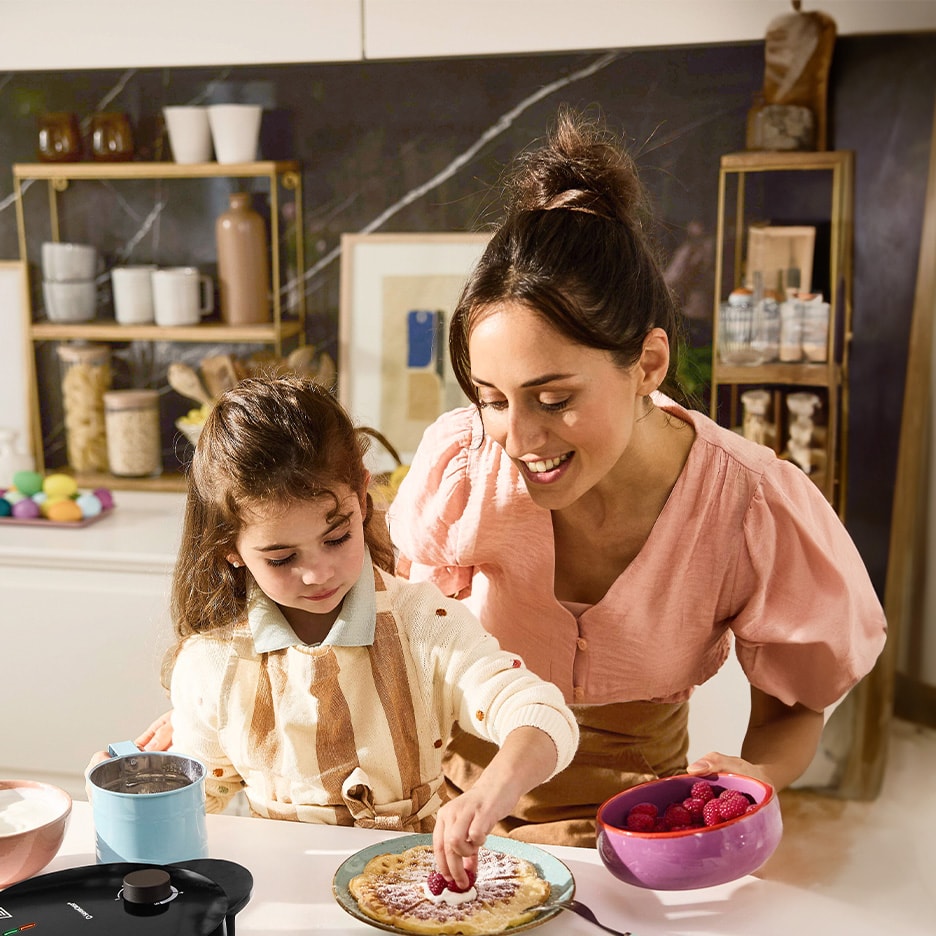 Mutter und Kind dekorieren Waffeln mit Himbeeren und Puderzucker in der Küche.
