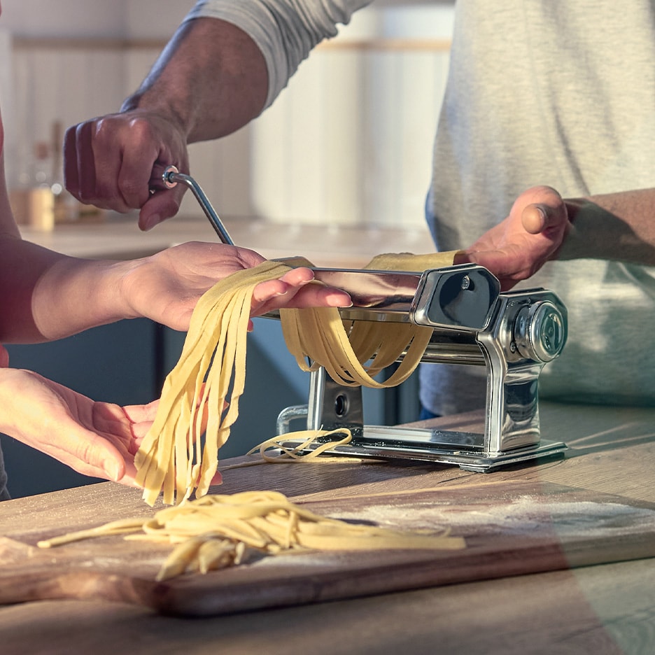 Hände, die eine Nudelmaschine benutzen, um frische Nudeln auf einem Holzschneidebrett zuzubereiten.