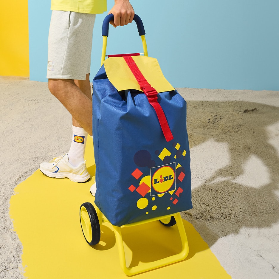 Blauer und gelber Einkaufstrolley mit Logo, auf einem gelben Laufsteg am Strand.