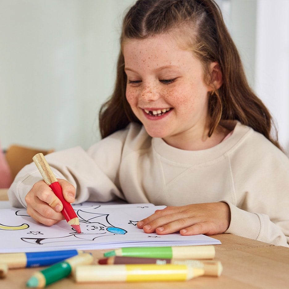 Lachendes Mädchen malt mit einem roten Buntstift in einem Malbuch.