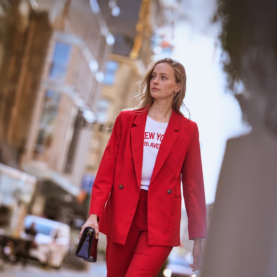 Frau in rotem Blazer und Hose, weißem T-Shirt, mit schwarzer Clutch, geht durch die Stadt.