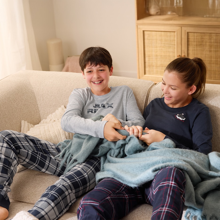 Zwei Kinder im Schlafanzug auf einem Sofa, teilen eine blaue Decke und lachen.