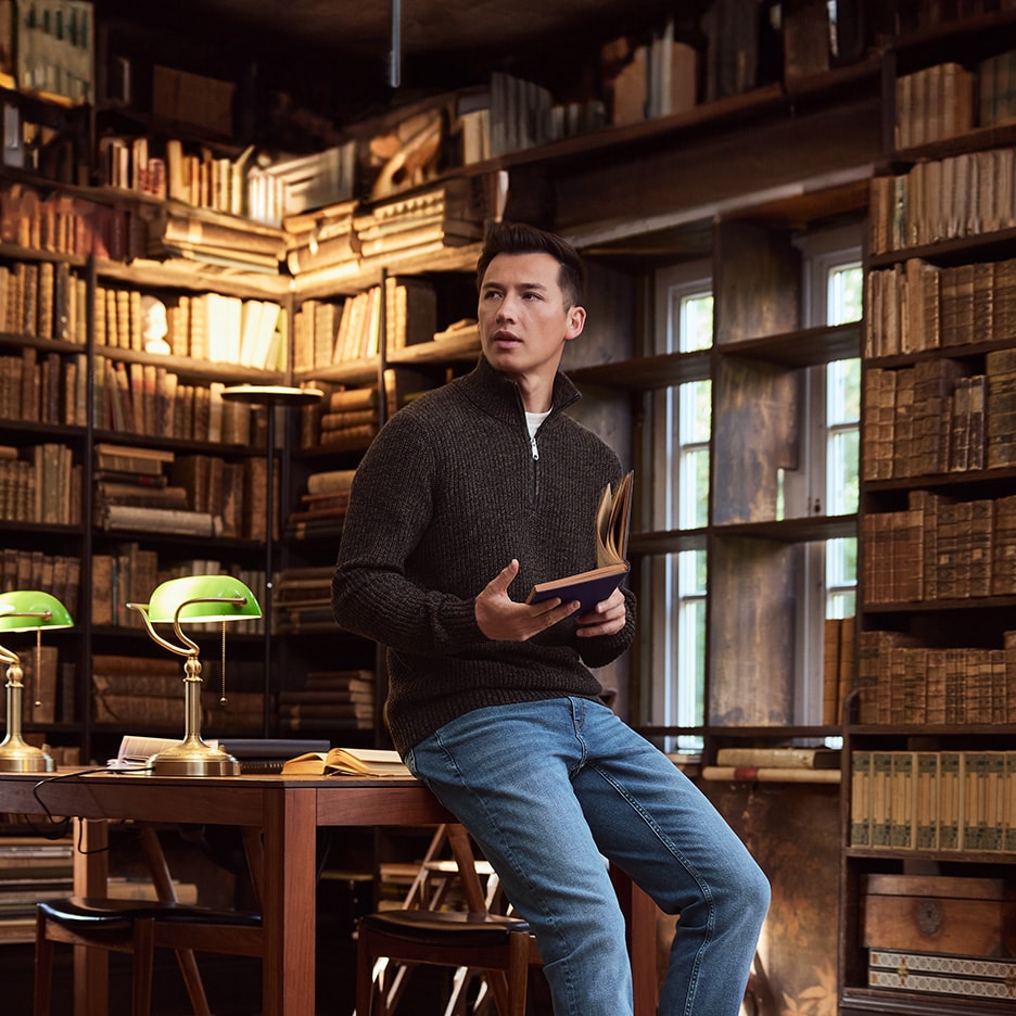 Mann in dunkelbraunem Half-Zip-Pullover und blauen Jeans, hält ein Buch in einer Bibliothek.