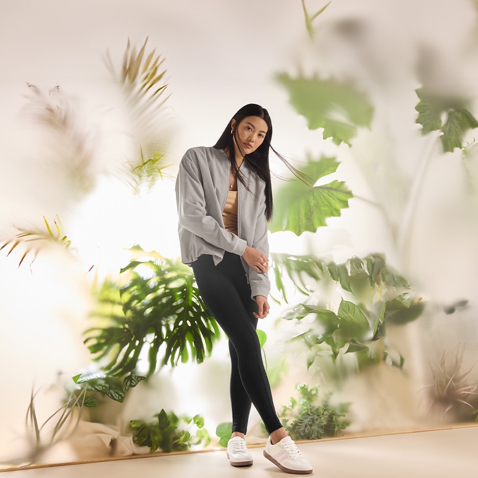 Frau in grauer Jacke und schwarzen Leggings vor Pflanzenhintergrund.