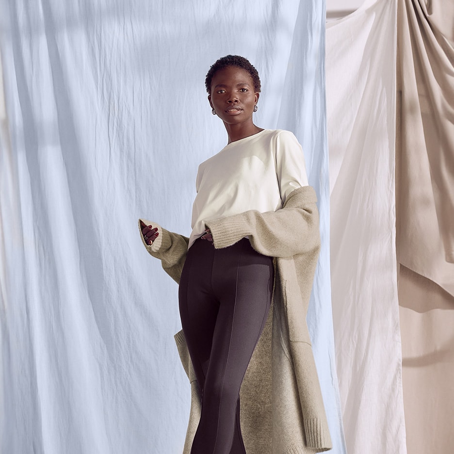 Femme en t-shirt blanc à manches longues, legging noir et cardigan beige devant des rideaux bleus et beiges.