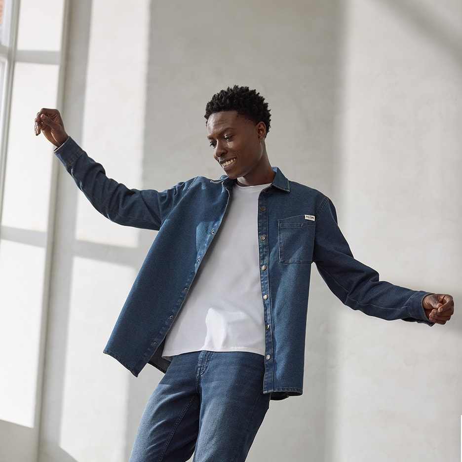 Un homme souriant en chemise en jean et t-shirt blanc, les bras levés.