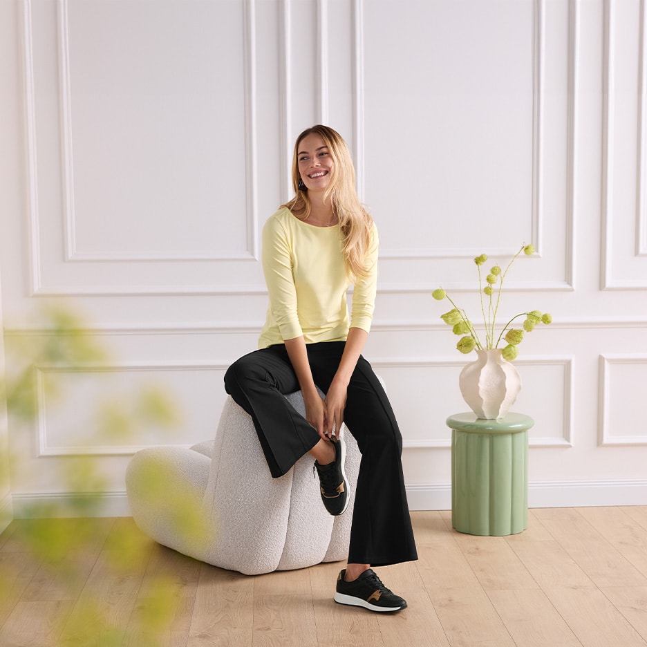 Lächelnde Frau in gelbem T-Shirt und schwarzer Hose sitzt auf einem Pouf.