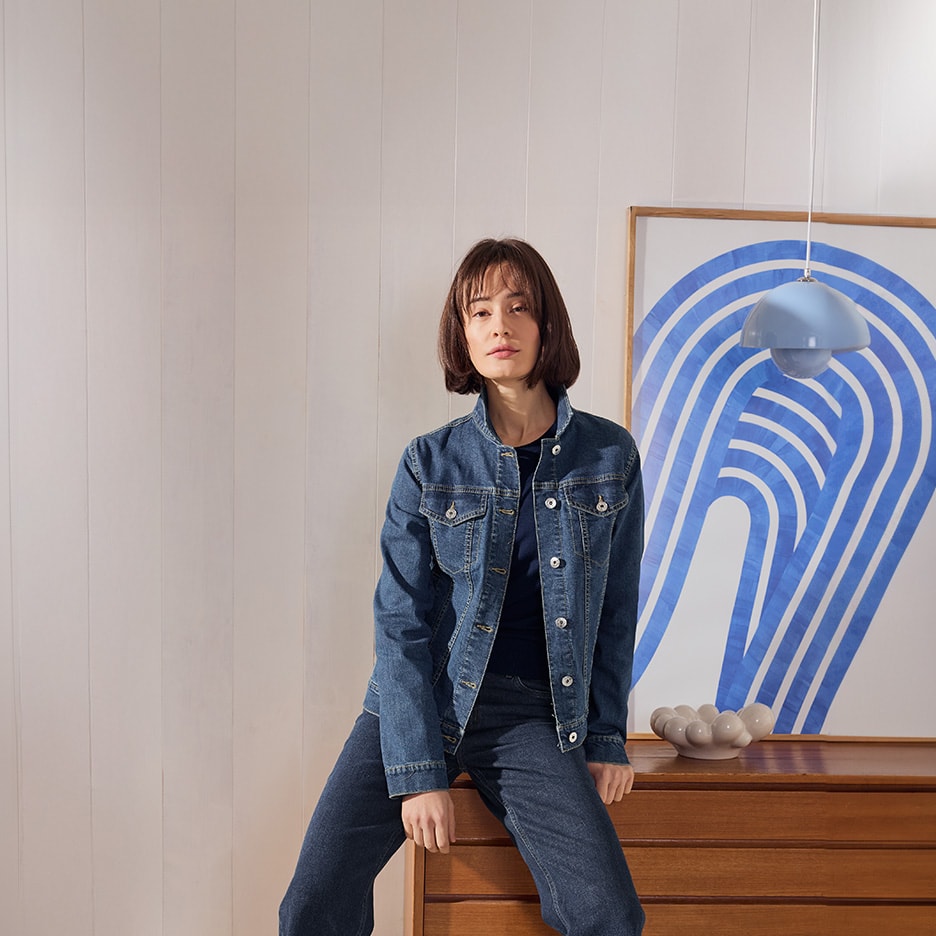 Femme en veste et jean en denim, assise sur une commode en bois avec un tableau abstrait bleu.